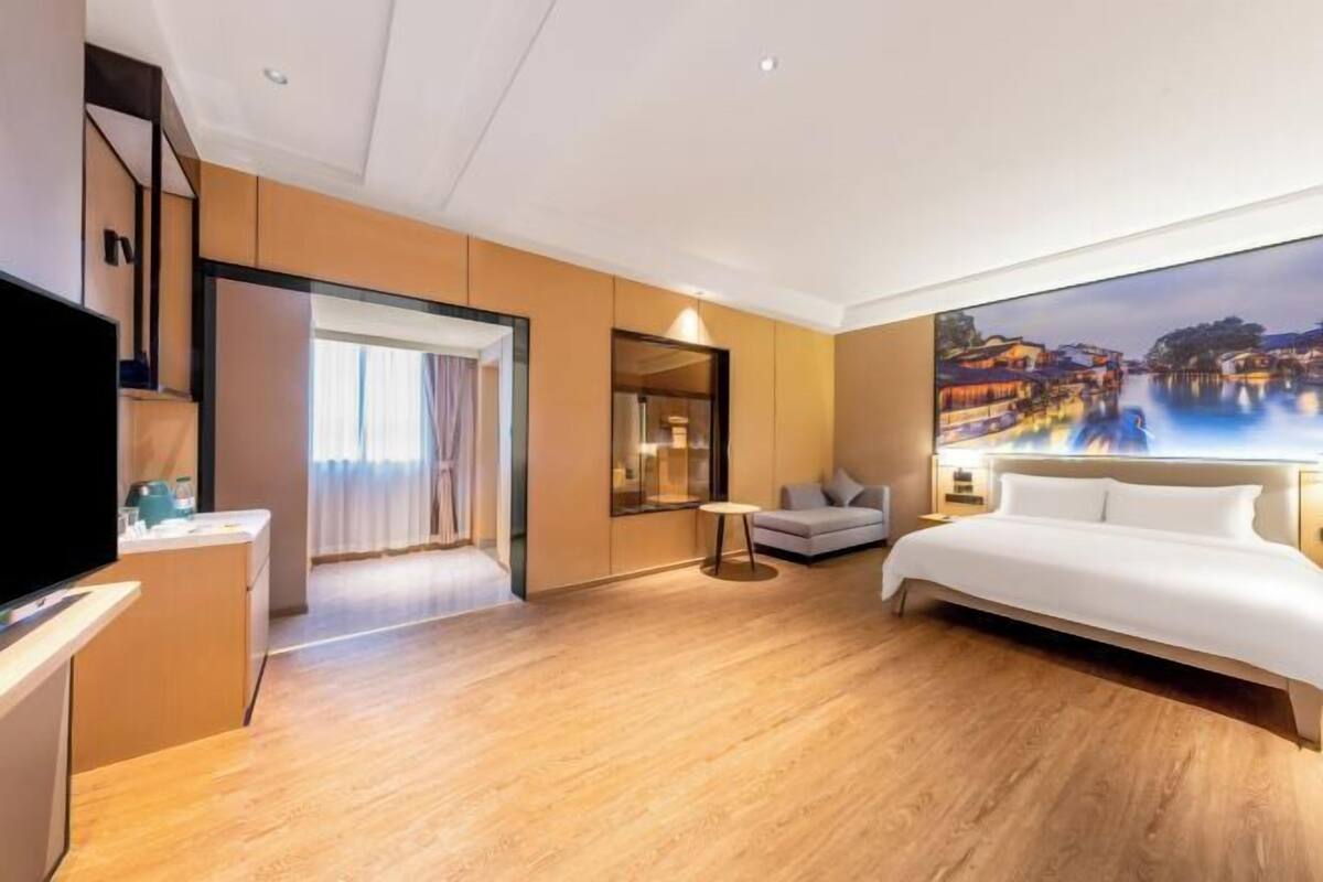 A hotel room with a bed, a TV, a desk, and a large window with a view of a water body.
