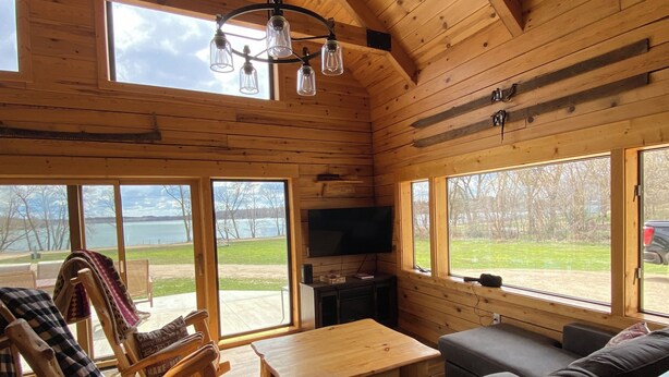 A cozy cabin interior with wooden walls, a large window offering a view of the outdoors, a television, and a seating area with a sofa and a rocking chair.