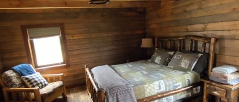A cozy log cabin bedroom with a wooden bed, a chair, a small table, and a window.