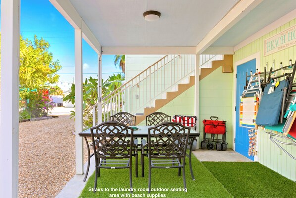 A covered outdoor seating area with a table and chairs, a staircase leading to an upper level, and a wall with beach supplies.