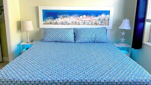 Ein Bett mit blauer Bettdecke, zwei passende Kissen, ein Nachttisch mit Lampe und ein Bild einer Strandansicht an der Wand.