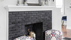 Una sala de estar moderna con chimenea de ladrillo negro, repisa blanca y objetos decorativos. Hay dos otomanes estampados y un sillón azul.