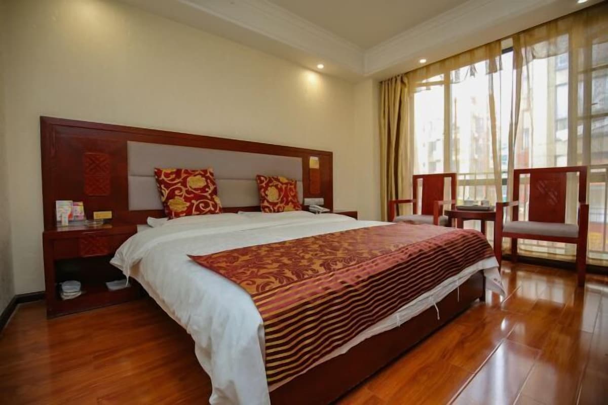 A hotel room with a large bed, wooden headboard, two nightstands, a chair, and a large window with curtains.