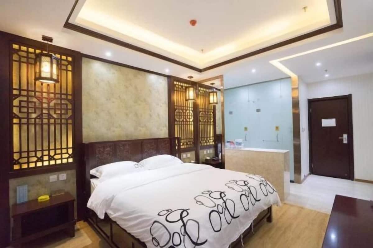 A modern hotel room with a bed, a nightstand, a wooden partition with decorative panels, and a bathroom visible through a glass door.