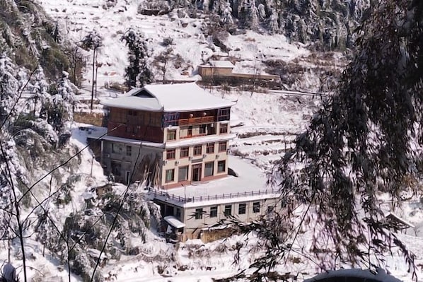 A multi-story building with a snow-covered roof, surrounded by trees and a mountainous landscape.