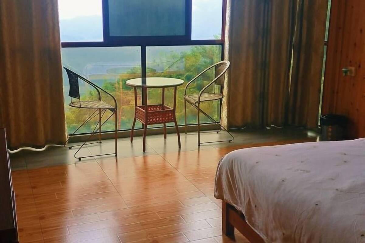 Een kamer met een bed, een klein tafeltje, twee stoelen en een groot raam met uitzicht op groen.