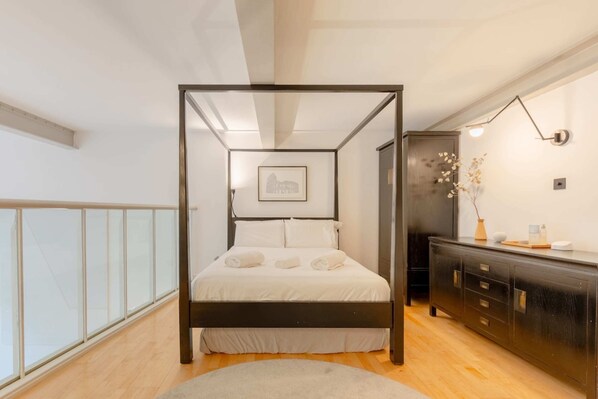 A modern bedroom with a four-poster bed, a wooden floor, a dark dresser, and a framed picture on the wall.