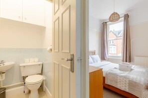 A bathroom with a toilet, sink, and a door leading to a bedroom with a bed, window, and curtains. - Roomy 3BD by Hampstead Heath! (London)