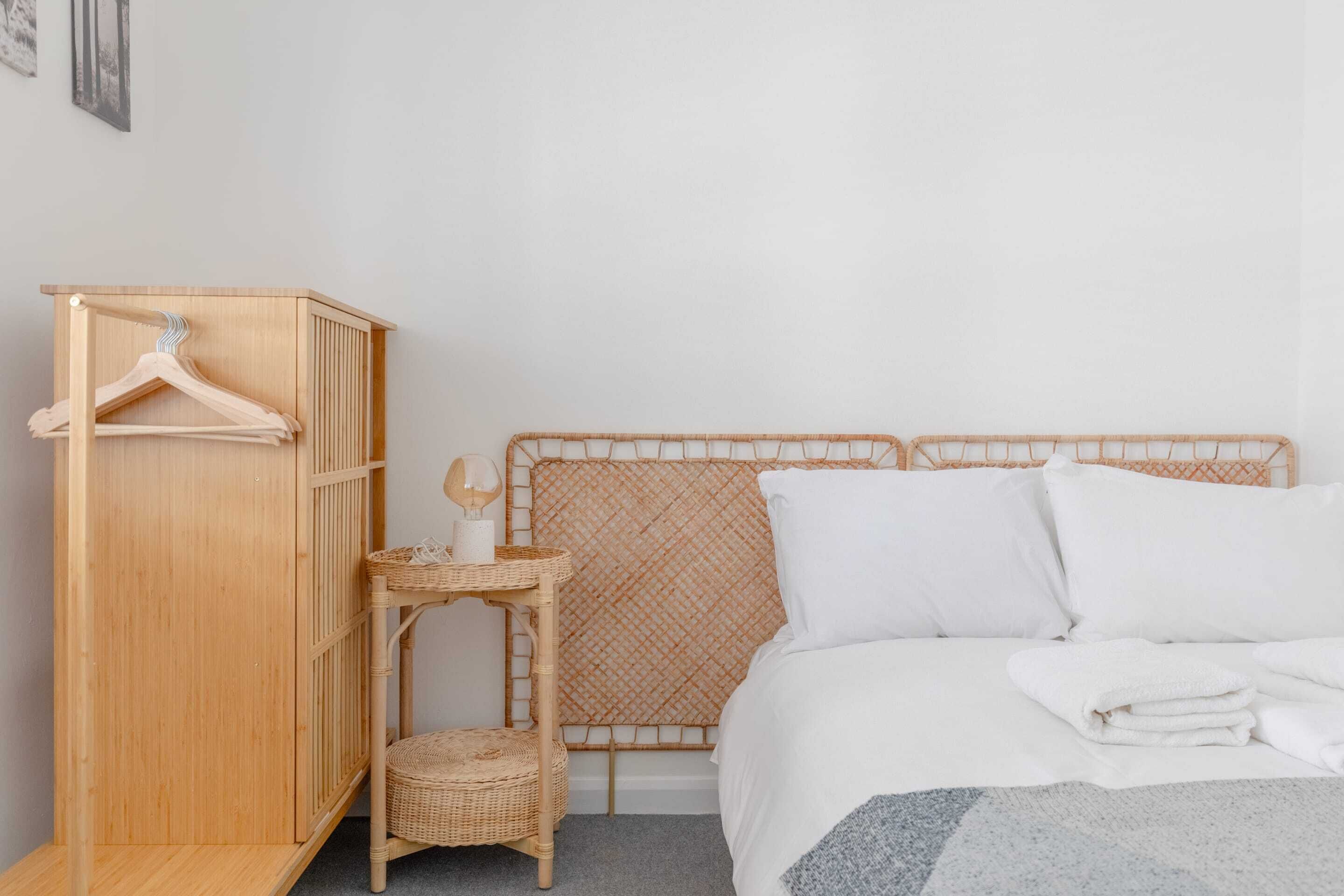 A bedroom with a wooden wardrobe, a wicker side table with a lamp, and a bed with white bedding.
