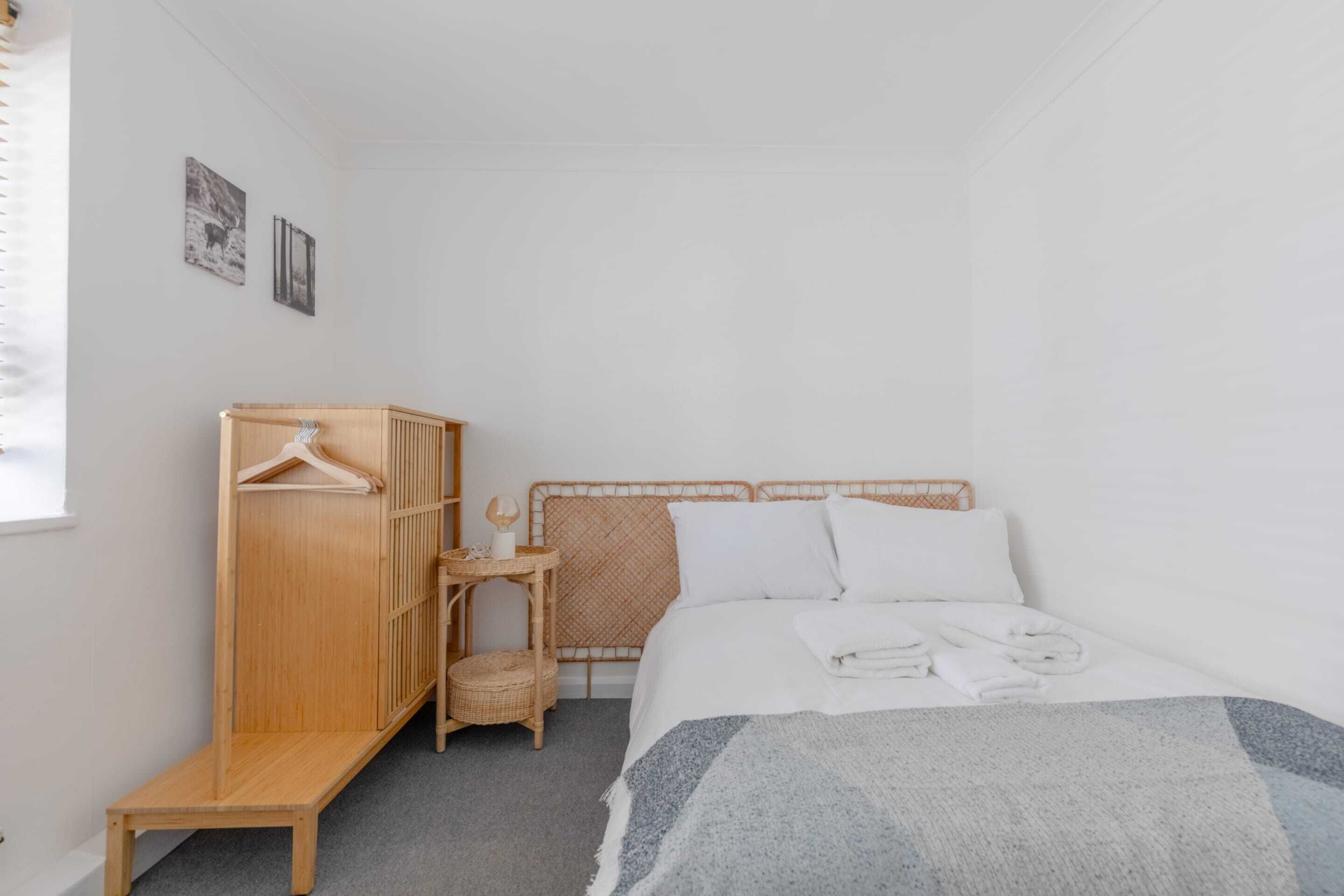 A bedroom with a bed, a wooden wardrobe, a small table, and a wall-mounted shelf.
