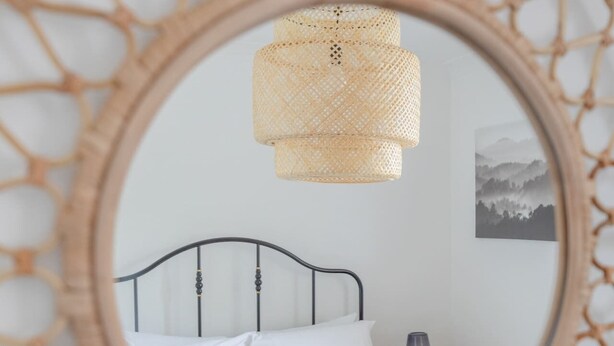A bed with white linens and a green towel, a bedside lamp, and a wall-mounted mirror with a woven frame. - Spacious 3BD Flat, Close to Tube - Fulham! (London)