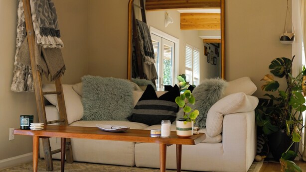 Ein gemütliches Wohnzimmer mit Holzboden, einer Couch mit Kissen, einem Couchtisch, einer an der Wand stehenden Leiter, einem Spiegel und einer Topfpflanze.