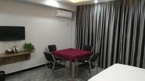 Une chambre d’hôtel dotée d’un téléviseur à écran plat fixé au mur, d’une étagère en bois avec une plante et une bouilloire, d’une table avec quatre chaises, d’un lit et d’une grande fenêtre avec des rideaux.