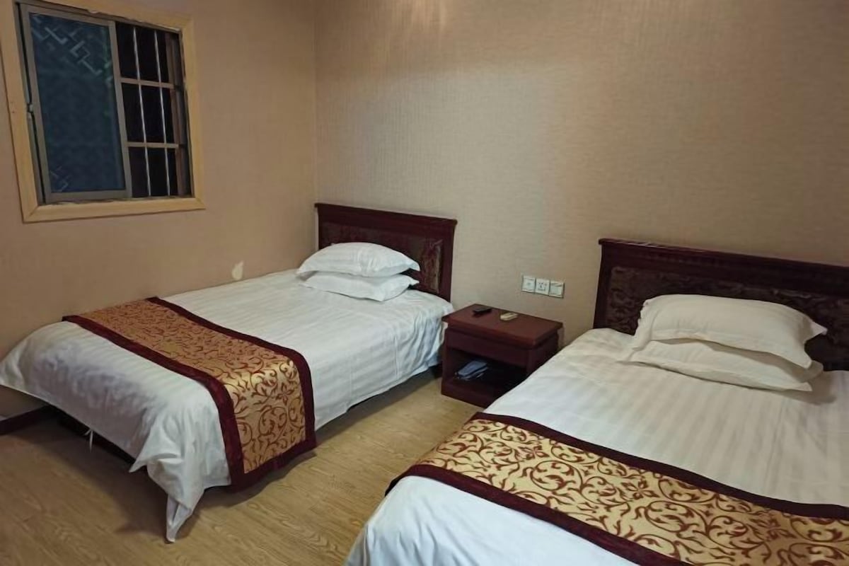 A hotel room with two single beds, wooden headboards, a bedside table with a lamp, and a window with bars.
