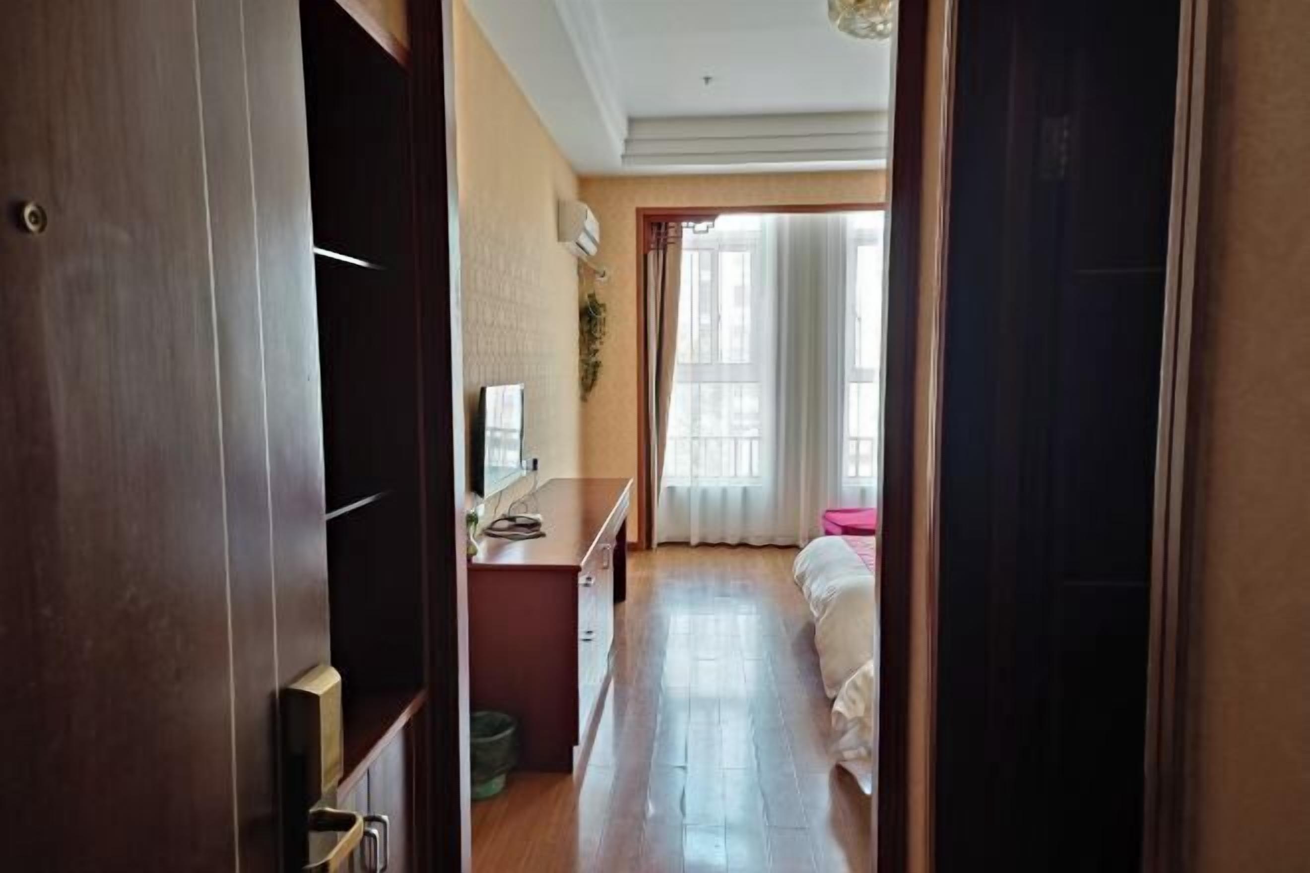 A hallway leading to a bedroom with a bed, a desk, and a window with curtains.