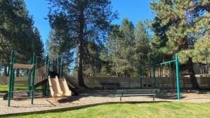 Une aire de jeux avec un toboggan, des balançoires et des bancs, le tout entouré d’arbres.