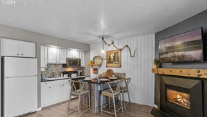 Une cuisine moderne avec des meubles blancs, un coin repas doté d’une table et de chaises en bois, une cheminée et un téléviseur fixé au mur.