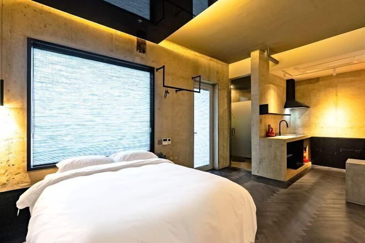 Una habitación de hotel moderna con una cama grande, vistas al agua, una kitchenette y una pared de concreto.