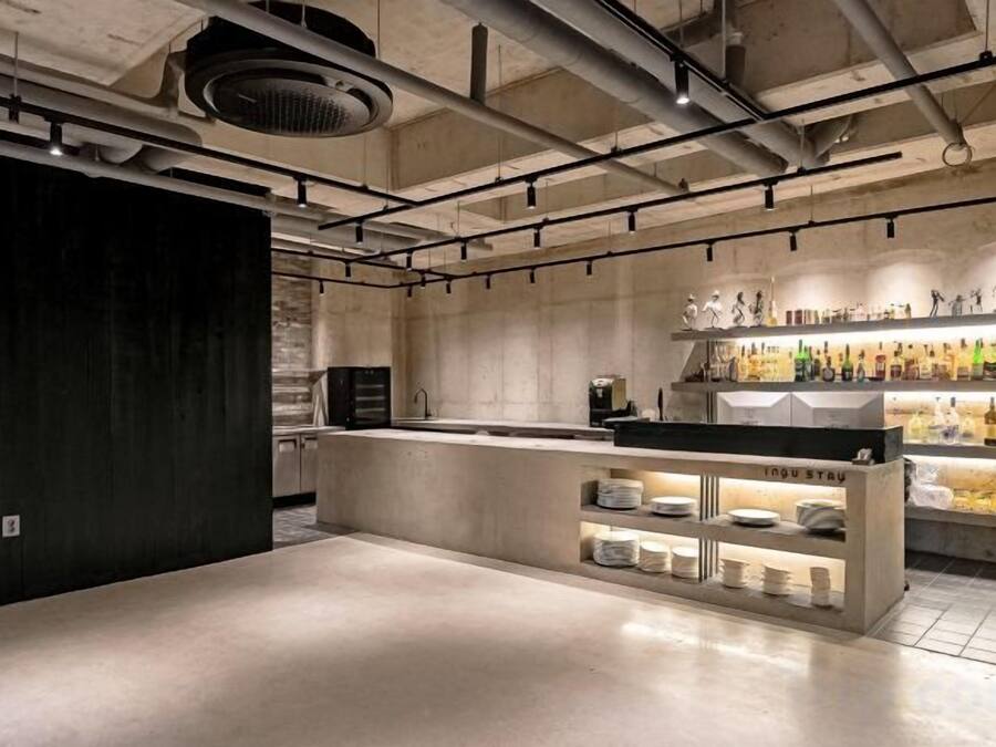 Zona de bar moderna con mostrador de hormigón, estanterías con botellas y sistema de ventilación en el techo.