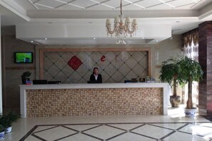 A hotel reception area with a receptionist, a television on the wall, and a large potted plant.