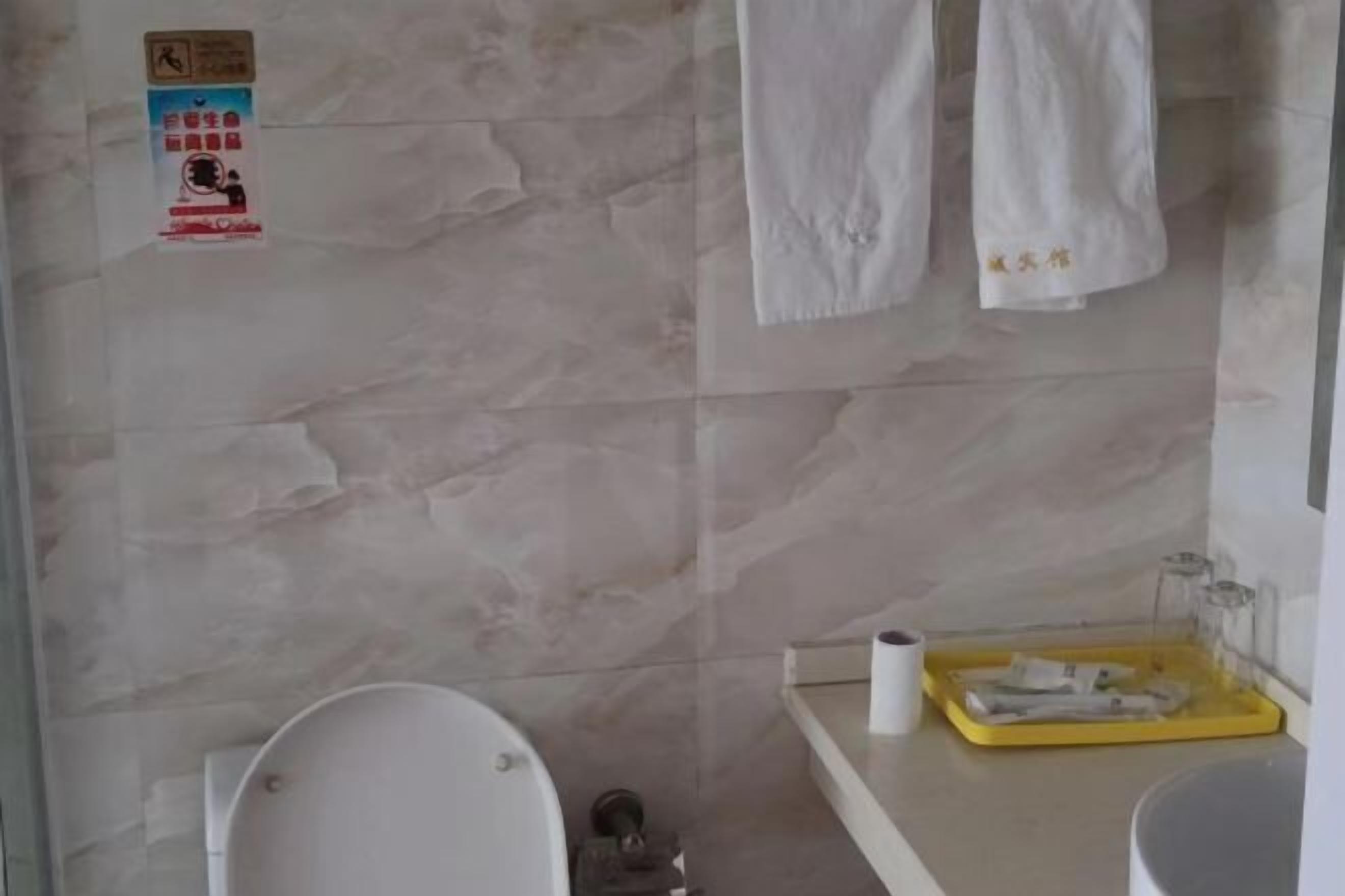 Un baño con inodoro, lavabo y un perchero para toallas. Hay un cartel en la pared y una bandeja amarilla con artículos de aseo.