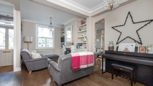 A living room with a piano, grey sofas, a star decoration on the wall, and a chandelier. - 3-bedroom Home in Richmond - Ideal Family Living (London)