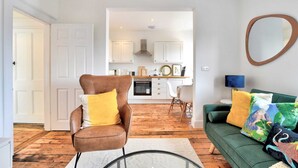 A modern living room with a green sofa, a brown leather chair, and a coffee table. A kitchen is visible in the background with white cabinets and a stove. - Stunning Apartment in the Heart of Chelmsford (Chelmsford)