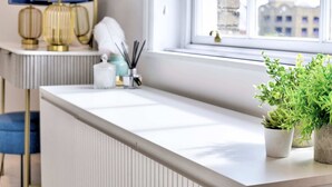 A modern bathroom with a white countertop, a window with a view of buildings, a blue chair, and a plant. - Stunning Apartment in the Heart of Chelmsford (Chelmsford)