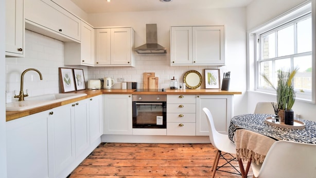 A modern kitchen with white cabinetry, a wooden countertop, and a built-in oven. There is a dining area with a table and chairs, and a window providing natural light. - Stunning Apartment in the Heart of Chelmsford (Chelmsford)