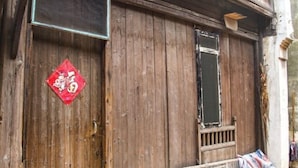 Un edificio rústico de madera con fachada desgastada, una ventana pequeña y un detalle decorativo rojo.