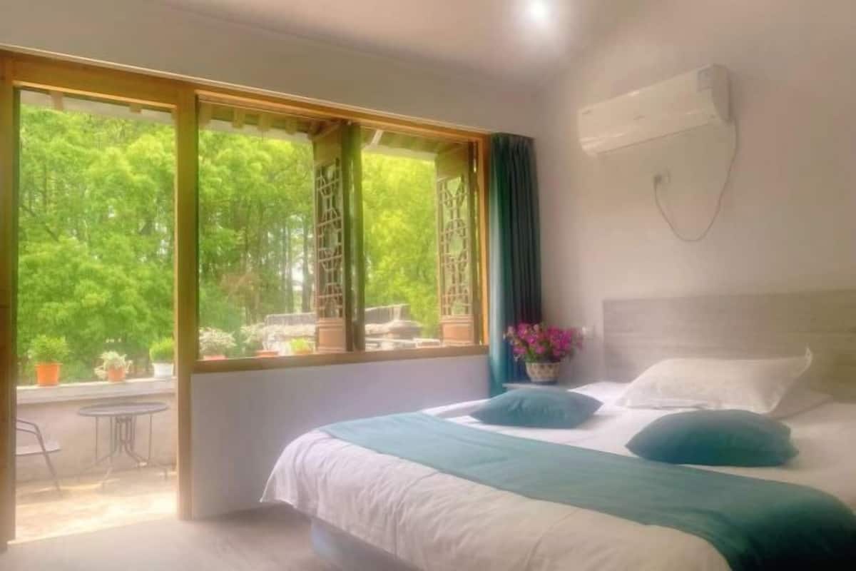 A bedroom with a bed, a window with a view of greenery, a table, and a chair outside.