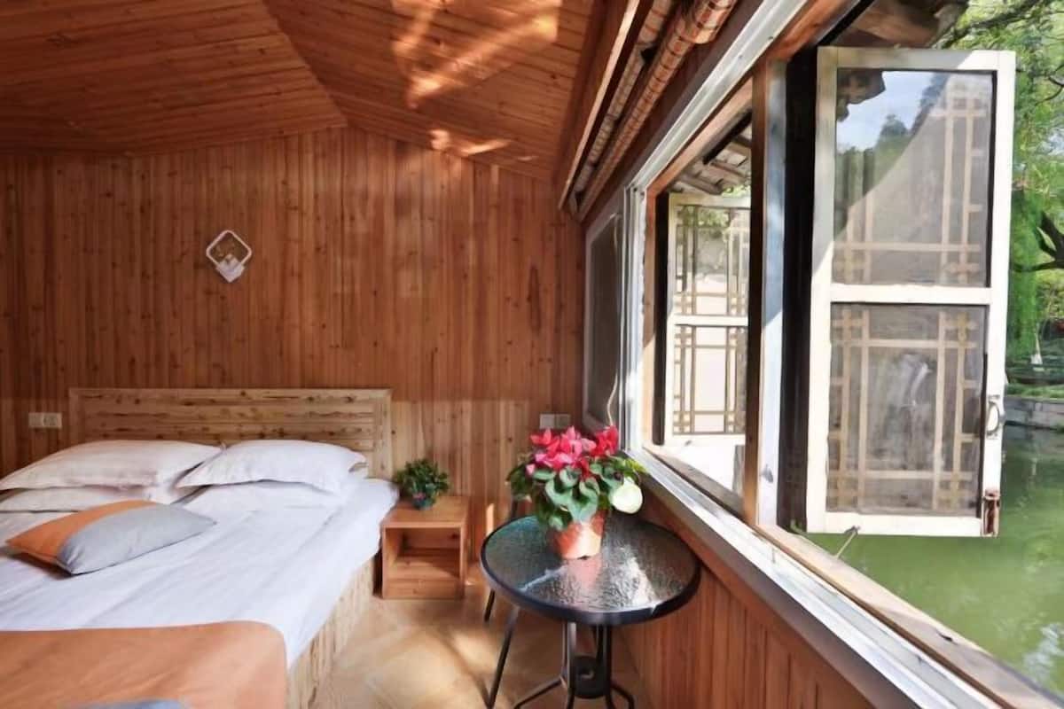 A wooden room with a bed, a small table with a plant, and a window with a view of greenery.