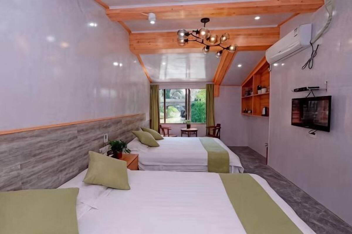 A hotel room with two beds, a TV, a small table, and a window with a view of greenery.
