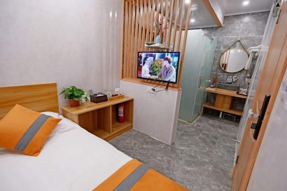 A hotel room with a bed, a TV, a small table with a plant, a shelf with toiletries, and a bathroom visible through an open door.