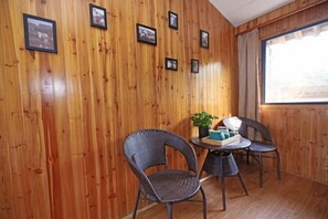 Une pièce aux murs en bois, ornée de tableaux encadrés, avec deux fauteuils en osier, une petite table ronde sur laquelle sont posés une plante et un plateau, et une fenêtre avec des rideaux.