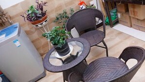 Un pequeño espacio al aire libre con sillas de mimbre y una mesa de vidrio, rodeado de plantas en macetas y un cubo de basura.