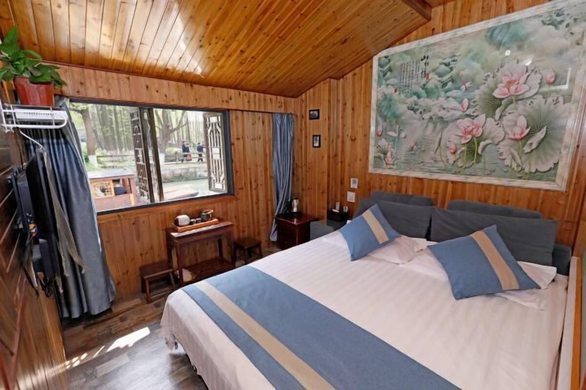 A wooden cabin room with a bed, a desk, a chair, a window, and a painting.