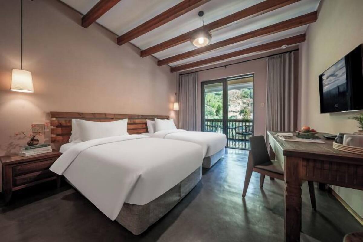 A hotel room with two beds, a wooden ceiling, a TV, a dining table, and a view of the outdoors.