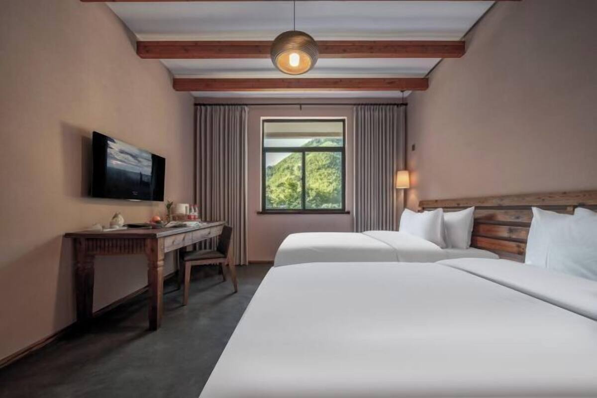A hotel room with a bed, a desk, a television, a window with curtains, and a wooden ceiling.