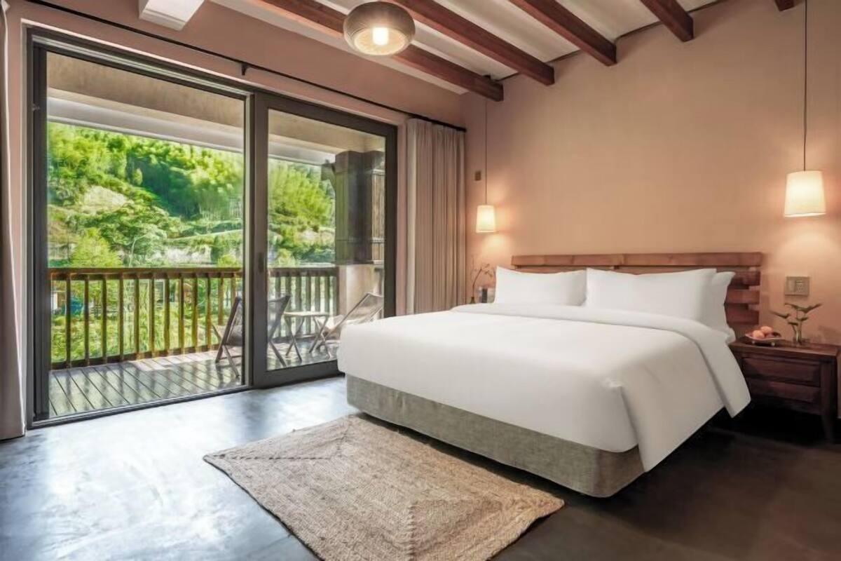 A bedroom with a large bed, a view through a sliding glass door to a balcony, and a wooden ceiling.