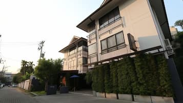 Un edificio moderno de dos pisos con techo plano, ventanas amplias y un balcón. Cuenta con un camino de entrada pavimentado y un seto a lo largo de un lateral del edificio.