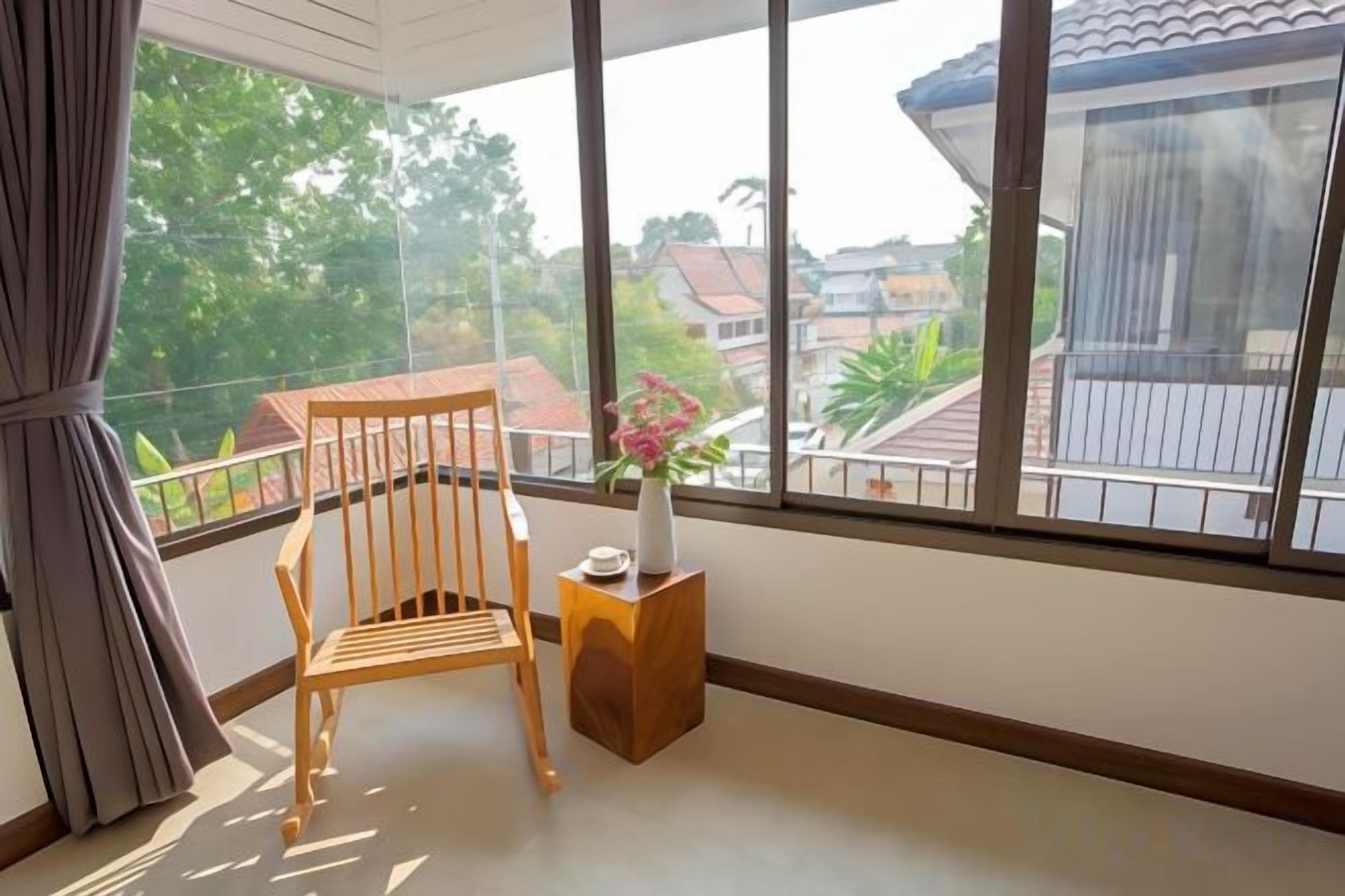 A room with a wooden chair, a small table with a vase, and a large window offering a view of buildings and trees.
