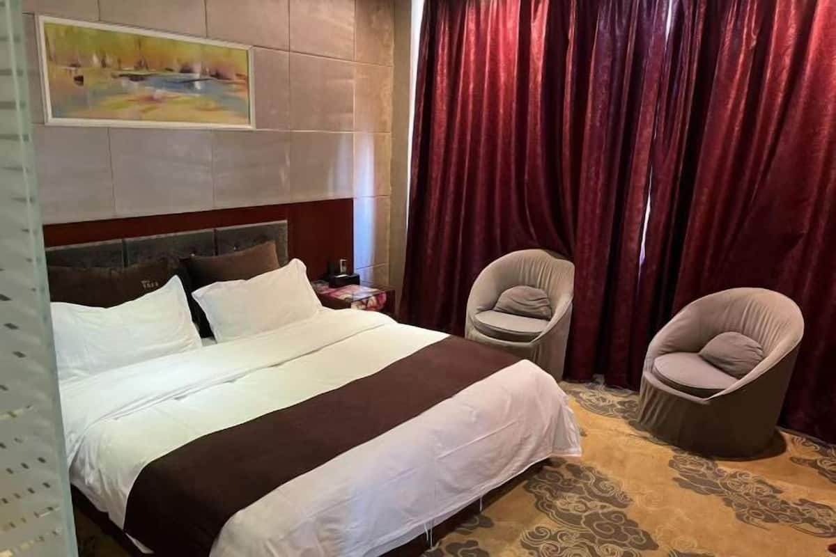 Een hotelkamer met een groot bed, twee fauteuils, een schilderij aan de muur en een rood gordijn.