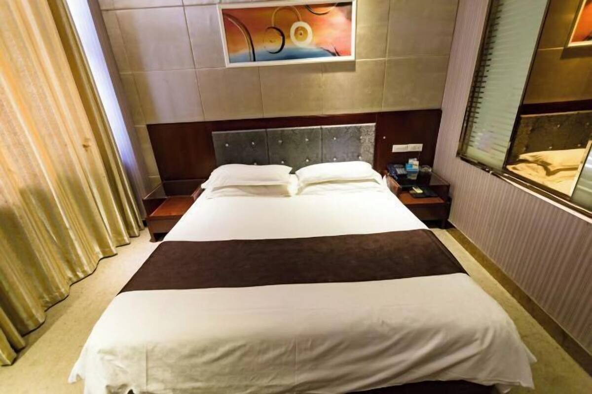 Een hotelkamer met een groot bed, een hoofdeinde, een nachtkastje, een schilderij aan de muur en een raam met gordijnen.