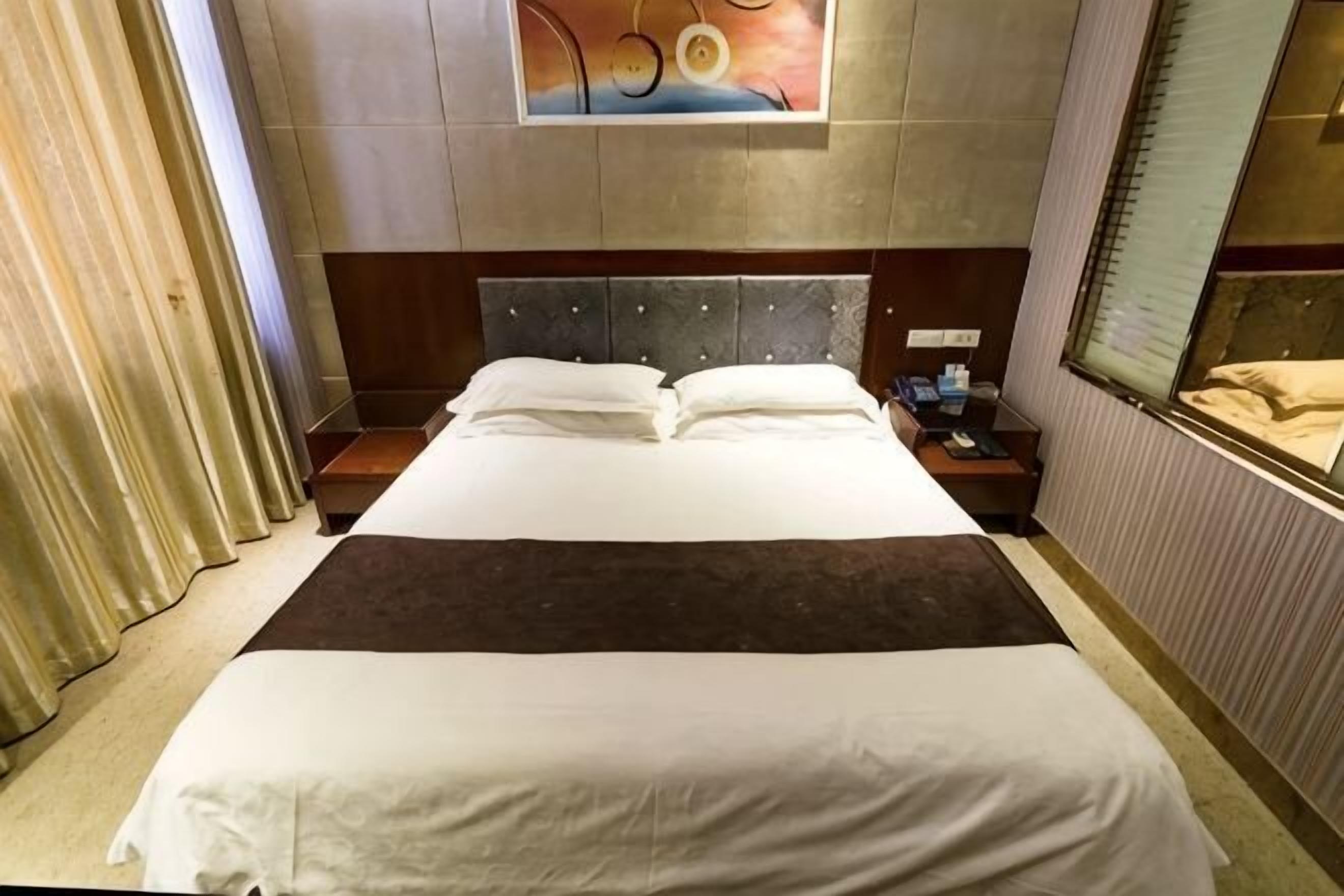 Hotellihuone, jossa on suuri sänky, sängynpääty, yöpöytä, ikkuna ja taulu seinällä.