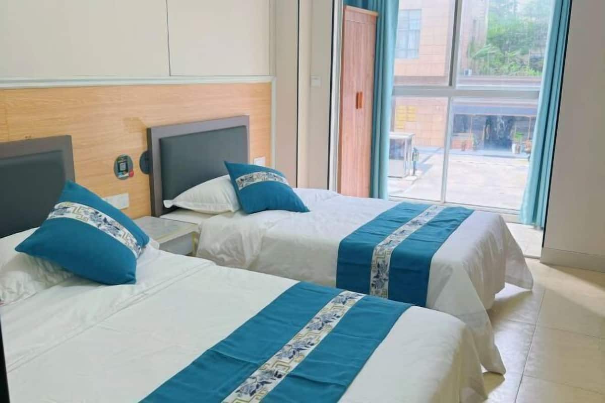A hotel room with two beds, each with a blue and white bedspread, a wooden headboard, and a window with a view of the outside.