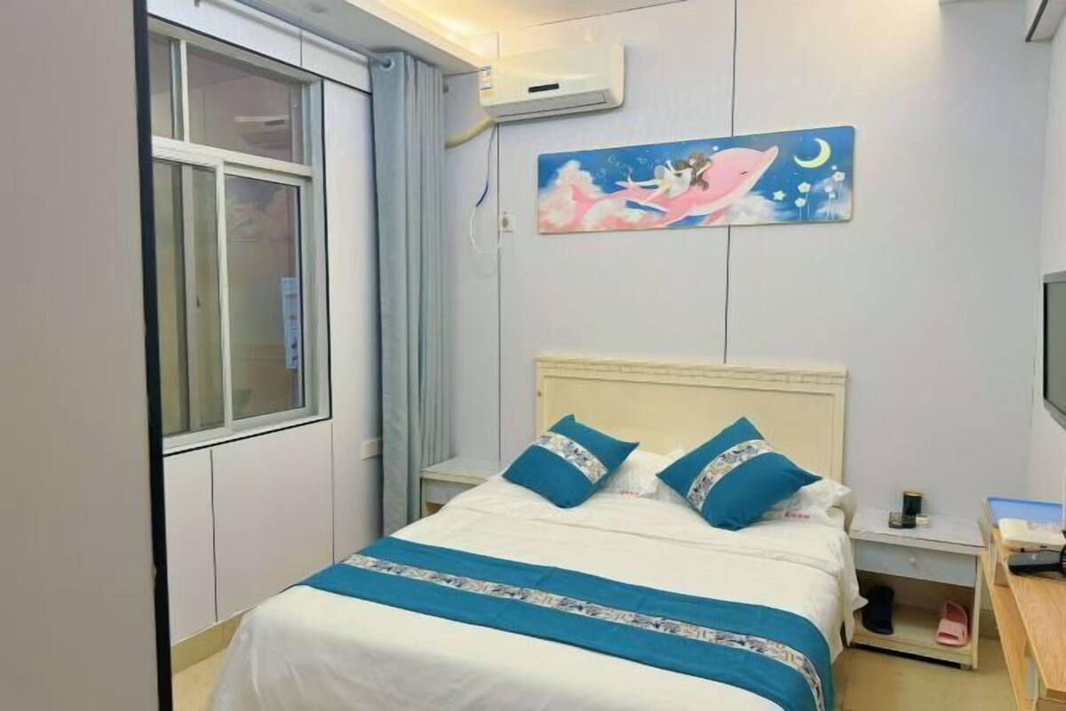A bedroom with a bed, a nightstand, a desk, a window, and a wall-mounted air conditioner.