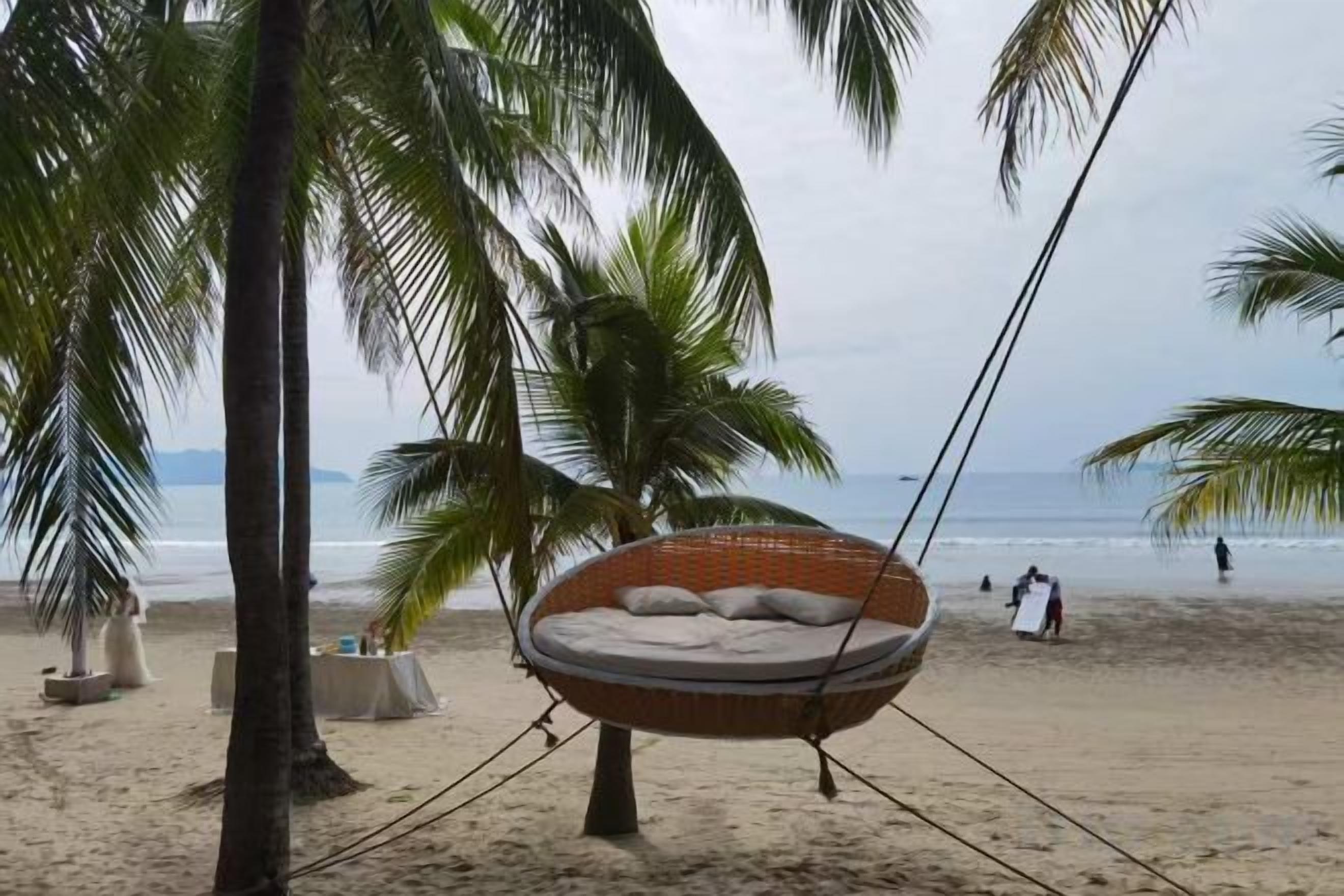 A beach setting with a hammock chair, palm trees, and a sandy beach.