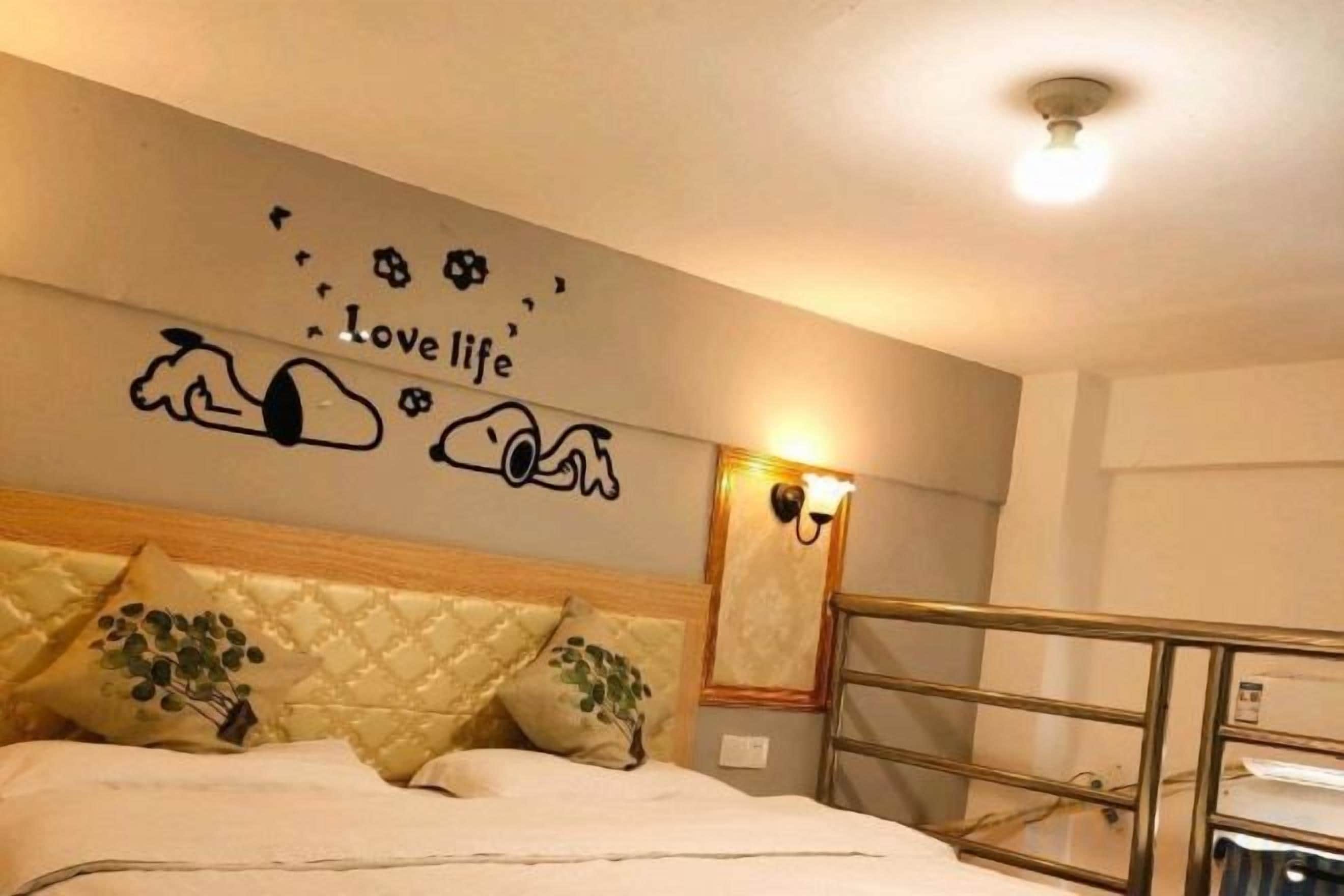 A hotel room with a bed, a headboard, a wall with a decorative phrase, and a light fixture.
