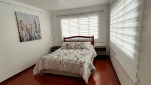 Un dormitorio con una cama, una mesita de noche, un estante en la pared y una ventana con persianas.
